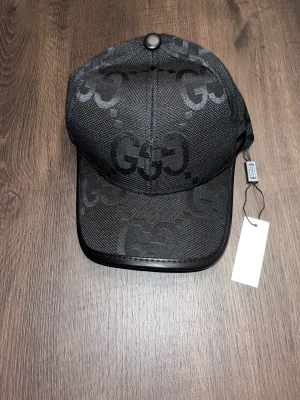 Svart keps med GG-mönster - Snygg svart keps med det klassiska GG-mönstret över hela kepsen. Kepsen har en böjd skärm med läderdetaljer och justerbar rem baktill i skinn. Materialet är polyester och skinn, och designen är riktigt stilren och lyxig.