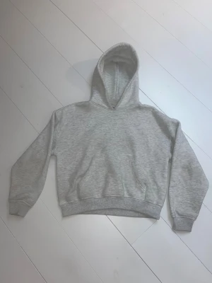 Grå hoodie med huva och ficka - Snygg grå hoodie med klassisk huva och stor magficka. Tröjan har ribbade muddar vid ärmslut och nederkant, vilket ger en skön och avslappnad passform. Perfekt för chill dagar och enkel att matcha med jeans eller mjukisbyxor.