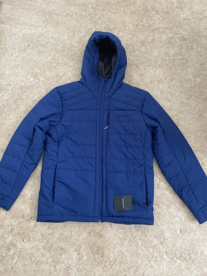 Blå pufferjacka Peak Performance XL - Blå pufferjacka från Peak Performance i storlek XL. Jackan har huva, dragkedja framtill och en bröstficka med diskret logga. Tillverkad i polyester och har quiltad design för extra värme. Perfekt för kyliga dagar när du vill hålla stilen.