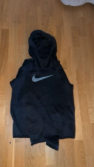 Svart Nike Dri-Fit hoodie utan ärmar - Svart hoodie från Nike med Dri-Fit-teknologi, perfekt för träning. Tröjan har huva, stor silverfärgad swoosh på bröstet och är ärmlös för extra rörelsefrihet. Materialet är syntet och designen är sportig och clean.