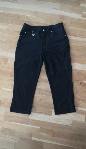 Svarta byxor från Röhnisch - Svarta byxor från Röhnisch med raka ben och praktiska fickor, inklusive en bakficka med dragkedja. Byxorna har bälteshällor och silverfärgade detaljer.