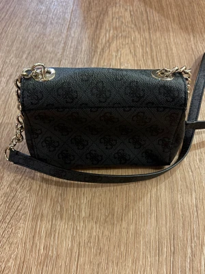 Svart väska från Guess  - I princip inga tecken på användning💓Snygg svart axelväska från Guess med subtilt mönster och guldfärgad kedja. Väskan har ett dekorativt lås framtill med Guess-logga och flera fack inuti, inklusive dragkedja. Perfekt storlek för det viktigaste och stilren design.