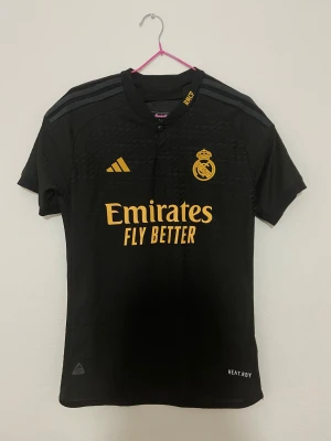 Svart Real Madrid fotbollströja Adidas - Snygg svart Real Madrid fotbollströja från Adidas med guldfärgade detaljer och logga. Tröjan har korta ärmar, rund hals och klassiska Adidas-ränder på axlarna. Tillverkad i lätt och ventilerande material med HEAT.RDY-teknologi. Perfekt för dig som älskar fotboll och vill ha en stilren look.