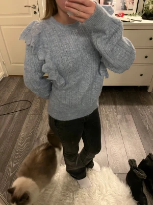 Ljusblå stickad tröja med volanger Lindex - Superfin ljusblå stickad tröja från Lindex med snygga volangdetaljer längs axlarna. Tröjan har ett diskret mönster, rund hals och långa ärmar med ribbade muddar. Perfekt för dig som gillar en söt och trendig look.