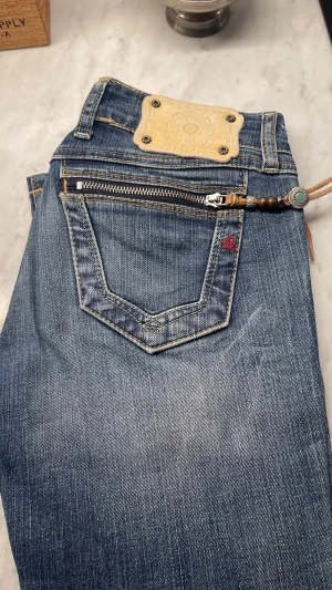 Blå Replay jeans med dragkedjedetalj - Snygga blå jeans från Replay med cool dragkedja och dekorativt hänge vid bakfickan. Klassisk denim med kontrastsömmar och läderpatch med blommönster. Perfekta för dig som vill sticka ut lite extra.