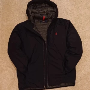 Mörkblå pufferjacka Polo Ralph Lauren - Snygg mörkblå pufferjacka från Polo Ralph Lauren med huva och röd logga på bröstet. Jackan har quiltad insida och har en stilren look som funkar till det mesta. Säljs då den inte används. Nypris ca 2700kr. Priset kan diskuteras