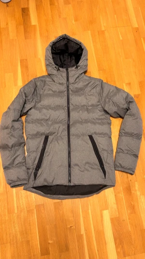 Grå pufferjacka från Everest - Grå pufferjacka från Everest med huva och svarta detaljer. Jackan har två snedställda fickor med dragkedja och en hel dragkedja framtill. Perfekt för kalla dagar med sin quiltade design och stilrena look.