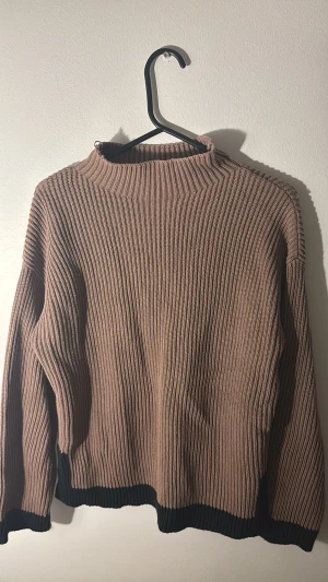 Ribbad brun stickad tröja från VILA - Säljer en ribbad stickad tröja från VILA i brun färg med svarta detaljer vid nederkant och ärmslut. Tröjan har lång ärm, hög hals och loose passform. Perfekt för dig som gillar enkel men snygg stil och vill ha något som är både bekvämt och trendigt. Finns liten missfärgning vid nederkant på tröjan men inget stort.