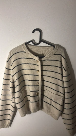 Beige randig kofta med guldknappar - Snygg beige kofta med svarta ränder och guldiga knappar framtill. Modellen är croppad med två fickor och har en rund halsringning. Stickad i mjukt material, perfekt för lager-på-lager. Passar dig som gillar klassisk men chill stil. Passar både XS och S