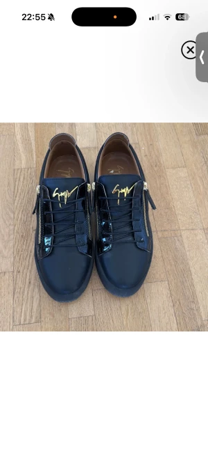 Svarta sneakers från Giuseppe Zanotti - Snygga svarta sneakers från Giuseppe Zanotti med guldfärgade dragkedjor och logga på plösen. Skorna har både matta och lackade partier i skinn, rund tå och svarta snören. Perfekta för dig som vill sticka ut med exklusiva detaljer.