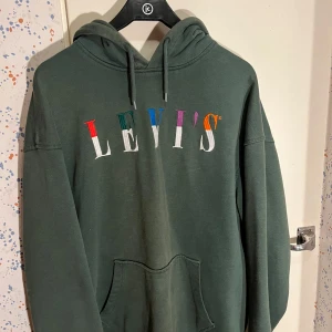 Grön hoodie från Levi's - Säljer en mörkgrön hoodie från Levi's med stor broderad logga i flera färger på bröstet. Tröjan har huva med snörning och en stor magficka framtill. Storlek M.