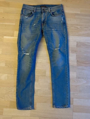 Blå jeans från Nudie med slitningar - Snygga blå jeans från Nudie med coola slitningar och distressed detaljer på både fram- och baksida. Modellen har rak passform och klassisk femficksdesign. Jeansen är tillverkade i bomull och har en normal midja. Perfekt för dig som gillar en avslappnad streetstil.