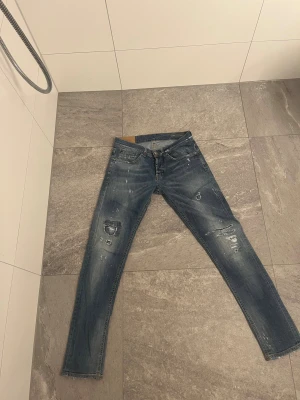 Blåa dondups jeans  - Snygga blå jeans med slitningar och hål på benen, perfekt för en cool streetstil. Modellen är gergoe  och har klassisk femficksdesign med knapp och dragkedja. Jeansen har en tvättad look och detaljer som ger dem en rå känsla. De är i w31.