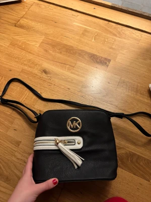 Svart axelväska - Snygg svart axelväska med vit dragkedjedetalj och tofs framtill. Väskan har MK-logga i guld och justerbar axelrem. Perfekt storlek för mobilen och det viktigaste. Stilren design som lyfter din outfit.