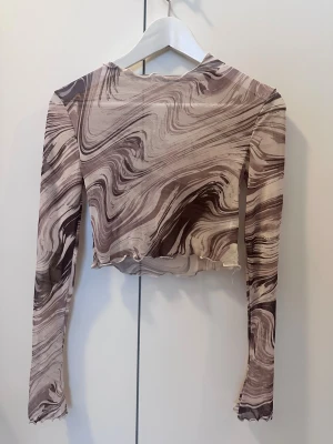 Brun mönstrad croppad långärmad topp - Säljer en croppad långärmad topp med unikt marmorerat mönster i olika nyanser av brunt och beige. Toppen har vågiga kanter vid ärmslut och nederkant för en cool touch. Perfekt för dig som gillar statement-plagg med snygg passform.