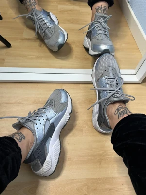 Nike Air Huarache silver/grå sneakers - Säljer ett par Nike Air Huarache sneakers i silver och grått med cool futuristisk vibe. Skorna har mesh och syntetpaneler, snörning och en chunky vit sula. Perfekta för dig som vill sticka ut med en sportig och modern look. Använda 3-4 gånger. Står att de är EU 41 men detta stämmer ej, de passar EU 39/25 cm. Köpta för 1200kr. Har lite smuts som eventuellt kan gå bort om de tvättas. 