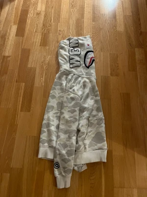 BAPE camo shark hoodie med dragkedja - Säljer en BAPE shark hoodie i ljus camo med ikonisk hajdesign på huvan och broderade WGM-bokstäver. Tröjan har hel dragkedja, ribbade muddar och fickor. Färgerna går i beige, grått och vitt. Perfekt för dig som gillar streetwear och vill sticka ut.