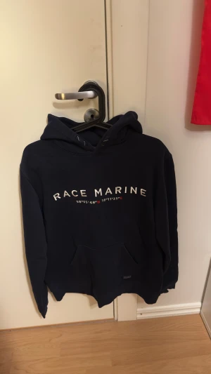 Mörkblå hoodie  - Snygg mörkblå hoodie från Race Marine i storlek M. Klassisk design med vit broderad logga och koordinater på bröstet. Stor känguruficka framtill och mjukt material som känns skönt mot huden. Perfekt för dig som gillar sportig och avslappnad stil.
