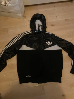 Svart Adidas vindjacka med huva - Svart och vit vindjacka från Adidas med klassiska tre ränder längs ärmarna, huva med snörning och dragkedja framtill. Jackan har Adidas-logga på bröstet och nedre delen, samt meshdetaljer i vitt. Perfekt för en sportig streetstil. Storlek är M men är stor i storlek. Jackan är i väldigt bra skick men är använd. Hängaren är av dock.