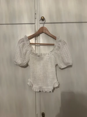 Vit blus med puffärm och volang - Säljer en söt vit blus med puffärmar och volangdetaljer vid nederkant och ärmslut. Blusen har en smockad kropp och är tillverkad i ett lätt, luftigt material med broderade detaljer. Perfekt för dig som gillar romantisk och feminin stil.