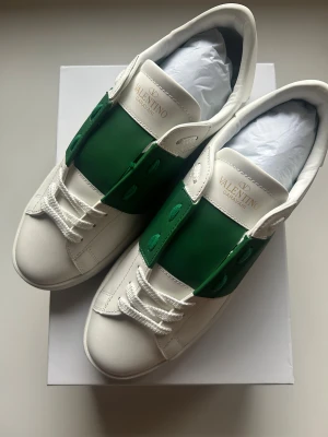Valentino Garavani Open sneakers vit/grön - Snygga Valentino Garavani Open sneakers i vitt skinn med bred grön läderpanel över mitten. Klassisk rund tå, platt sula med ikoniska nitar längs kanten och snörning framtill. Märkeslogga på plös och undersida. Perfekt för dig som gillar exklusiv streetstyle. OBS 1799kr utan box!