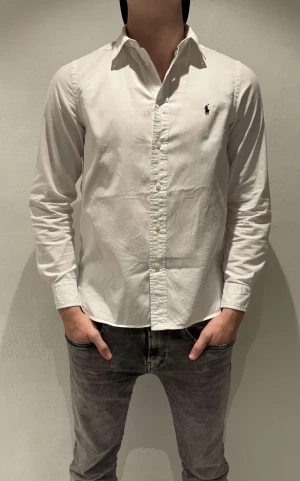 Vit skjorta från Ralph Lauren - Klassisk vit skjorta från Ralph Lauren med broderad logga på bröstet. Skjortan har lång ärm, knappar framtill och en snygg krage. Tillverkad i mjuk bomull och har en clean look som passar till allt.