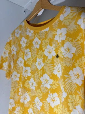 Gul blommig t-shirt från Morris - Fräsch gul t-shirt från Morris med vit blommigt mönster och tropiska blad över hela plagget. Rund hals och korta ärmar, perfekt för sommarens chill. Mjuk bomull som känns skön mot huden. Liten Morris-logga broderad på bröstet.