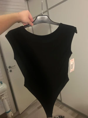 Svart bodysuit med öppen rygg - Säljer en svart bodysuit som är öppen i ryggen. Modellen har en tight passform och är tillverkad i mjukt, stretchigt syntetmaterial som sitter snyggt mot kroppen. Perfekt att styla med jeans eller kjol för en clean look. Endast testad men lapp kvar!