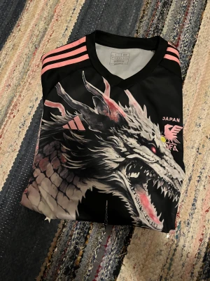 Japan Adidas fotbollströja drakmotiv - Säljer en svart Japan fotbollströja från Adidas med grymt drakmotiv och pagoda på framsidan. Tröjan har rosa detaljer och korta ärmar, samt tryck med körsbärsblommor. Materialet är lätt och andas, perfekt för match eller träning.