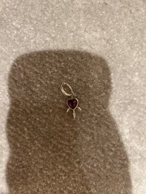 Örhänge med röd hjärtberlock - Coolt örhänge i silverfärgat metall med en röd hjärtformad berlock och tre spetsiga detaljer som sticker ut från hjärtat. Perfekt för dig som gillar edgy och unika smycken med lite attityd.