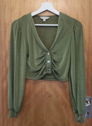 Olivgrön croppad blus från Gina Tricot - Säljer en olivgrön croppad blus från Asos med v-ringning och snygg rynkning framtill. Blusen har långa puffärmar och knappar i mitten. Mjukt och stretchigt material som sitter bekvämt och ger en cool siluett. Det finns lite slitage på toppen. 