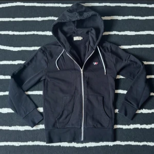 Svart hoodie från Maison kitsune - Svart hoodie från Maison kitsune med hel dragkedja, huva med vita snören och liten broderad logga på bröstet. Mjuk bomullskänsla och känguruficka framtill. Perfekt för chill dagar eller när du vill ha en enkel streetstyle. Kom med prisförslag!!