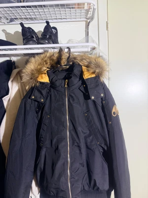 Svart pufferjacka från Moose Knuckles - Svart pufferjacka från Moose Knuckles med guldfärgade detaljer och logga på ärmen. Jackan har en stor huva med fluffig päls, dragkedja framtill och flera fickor med tryckknappar. Insidan av huvan har guldfärgat foder som sticker ut. Perfekt för kalla dagar. Storlek xxl men sitter som XL använt den 2 gånger bara pris är ej hugget i sten 