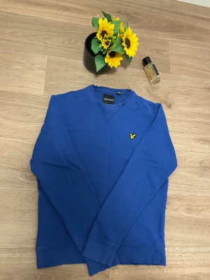 Blå sweatshirt från Lyle & Scott - Snygg blå sweatshirt från Lyle & Scott med klassisk gul logga på bröstet. Tröjan har rund hals, ribbade muddar och är långärmad. Perfekt för dig som gillar stilren design och vill ha en tröja som funkar till många olika outfits.