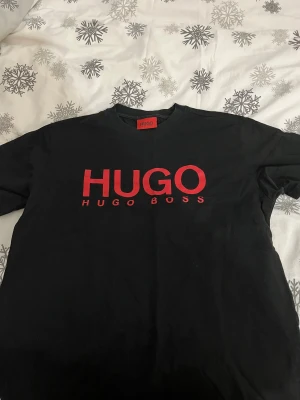 Svart t-shirt från Hugo Boss - Svart t-shirt från Hugo Boss med stor röd HUGO-logga på bröstet. Klassisk rund hals och korta ärmar. Materialet är mjuk bomull som känns skönt mot huden. Perfekt för dig som gillar stilrena och tydliga designer.