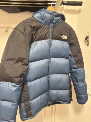 Blå dunjacka från The North Face - Säljer en blå och mörkgrå dunjacka från The North Face med huva. Jackan är quiltad och har en klassisk pufferstil som håller dig varm under kalla dagar. Perfekt för vintern och har en sportig look. Nypris 4000kr. Defekt enligt bild 2. Annars bra skick