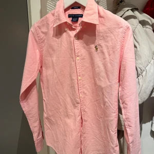 Rosa skjorta från Ralph Lauren - Snygg rosa skjorta från Ralph Lauren. Klassisk krage, långa ärmar och knappar hela vägen framtill. Ikoniska broderade loggan på bröstet ger en clean och stilren vibe. Perfekt för dig som gillar preppy och tidlös stil. Den passar någon som är 175 cm +- 5 cm