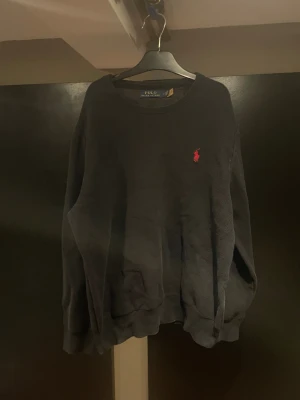 Svart tröja från Polo Ralph Lauren - Svart långärmad tröja från Polo Ralph Lauren med klassisk rund hals och röd broderad logga på bröstet. Tröjan har en loose passform och är tillverkad i mjuk bomull, perfekt för chill dagar. Snygg och enkel att matcha med jeans eller joggers.