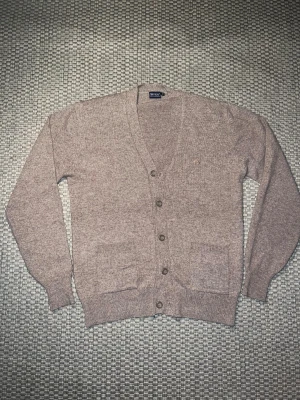 Beige kofta från To Wear - Klassisk beige kofta  med v-ringad hals och fem knappar framtill. Tröjan har två praktiska fickor och ribbade muddar vid ärmslut och nederkant. Perfekt för lager-på-lager och chill stil under höst och vinter.