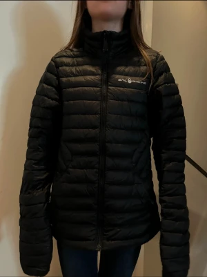 Svart dunjacka från Sail Racing - Säljer en svart dunjacka från Sail Racing med horisontella quiltade sektioner och hög krage. Jackan har dragkedja framtill och logga på bröstet. Perfekt för kalla dagar och har en clean, sportig look som funkar till jeans eller hoodie.