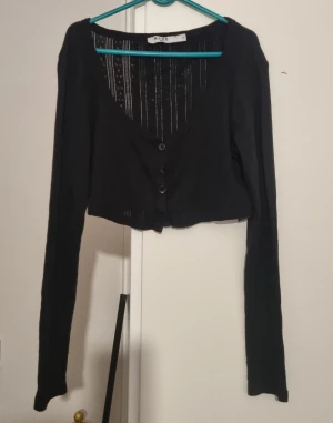 Svart croppad bolerotopp från NA-KD - Svart bolerotopp från NA-KD med croppad passform och lång ärm. Toppen har v-ringning, knappar framtill och ett diskret ribbat mönster med små hål och ränder på ryggen. Perfekt för att ge din outfit en cool layering-effekt.