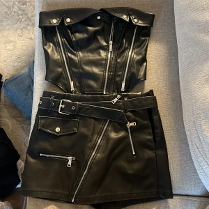 Svart skinnkjol med dragkedjor och bälte - svart kjol i skinnimitation från NewYorker med flera silvriga dragkedjor, bälte i midjan och fickdetaljer. Inbyggda shorts och matchande topp. Båda strl M