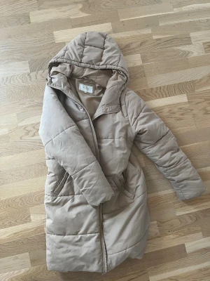 Beige lång pufferjacka med huva - Säljer en beige pufferjacka med huva och dragkedja framtill. Jackan är lång, har två sidofickor och quiltade sömmar som ger en schysst look. Perfekt för kalla dagar när man vill vara både varm och snygg. Passar dig som gillar en clean och enkel stil.