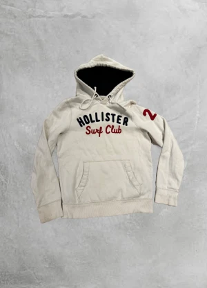 Vintage Hollister Hoodie🏄‍♂️ - Vit hoodie från Hollister med broderad text 'Surf Club' i rött och mörkblått på bröstet samt en röd siffra 2 på ärmen. Tröjan har snörning i huvan, känguruficka och mörkblått foder i huvan. Perfekt för chill dagar och streetstyle.