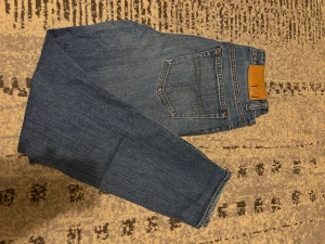 Tiger of Sweden jeans LEA blå 27/32 - Snygga blå jeans från Tiger of Sweden, modell LEA. Klassisk femficksdesign med bruna sömmar och läderpatch bak i midjan. Jeansen har rak passform och är tillverkade i slitstarkt bomullsmaterial. Perfekt för dig som gillar stilrena och tidlösa jeans.