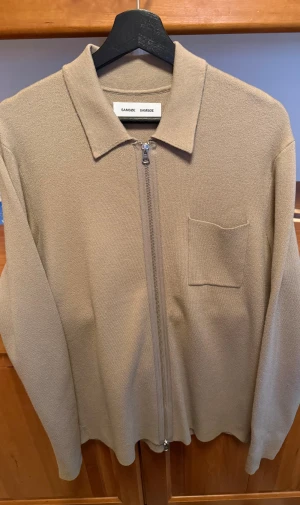 SAMSØE SAMSØE Guna Cardigan Full Zip - Snygg beige cardigan från Samsøe Samsøe med klassisk krage och hel tvåvägs dragkedja framtill. Tröjan har en enkel bröstficka och är tillverkad i ett mjukt material som ger en soft och stilren känsla. Perfekt för lager-på-lager. Aldrig använd, nytt skick.