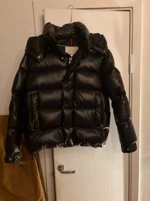 Moncler 70th Anniversary Maya Jacka - Säljer en svart dunjacka från Moncler med fet logga på ärmen och coolt serietryck på insidan. Jackan har hög krage, dragkedja och knappar, samt flera fickor med zip. Puffad design och glansigt material som ger en riktigt clean look. Priset går att diskutera.