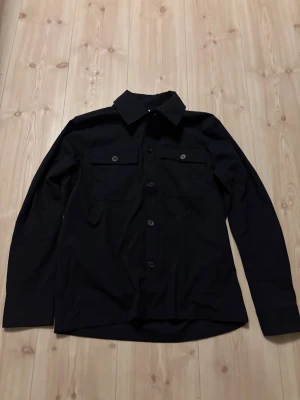 Blå selected homme overshirt  - Stilren blå overshirt med klassisk krage och fyra knappar framtill. Två stora bröstfickor med lock och knapp ger en cool utility-vibe. Långärmad modell i mjukt bomullstyg, perfekt att slänga över en t-shirt. Enkel och clean design som funkar till allt.