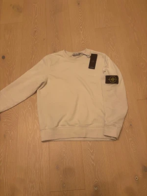 Vit sweatshirt från Stone Island - Riktig Stone Island. Vit sweatshirt från Stone Island med klassisk rund hals och ribbade muddar. Tröjan har den ikoniska Stone Island-loggan på vänster ärm och är tillverkad i mjuk bomull. Perfekt för dig som gillar clean och stilren streetwear.