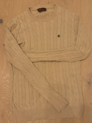 Beige kabelstickad tröja Morris M - Snygg beige kabelstickad tröja från Morris i 100% pima bomull. Tröjan har rund hals, lång ärm och ett broderat mörkt emblem på bröstet. Perfekt för dig som gillar klassisk stil med en modern twist.